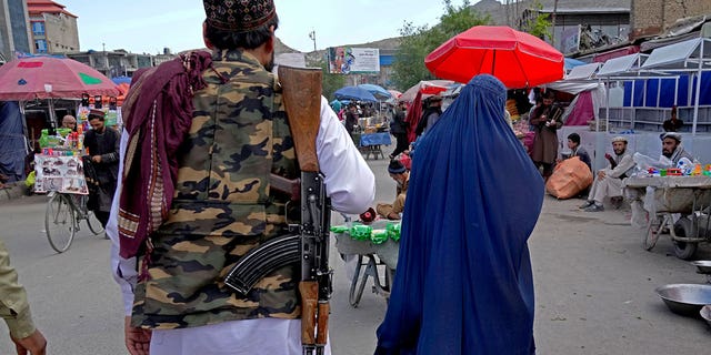 A woman wearing a burka walks through the old market as a Taliban fighter stands guard, in downtown Kabul, Afghanistan, Sunday, May 8, 2022. Afghan women are furious and fearful over a recent decree by the country's Taliban leaders that reinstated the burqa and similar outfits as mandatory for them in public. But some are defying the strict interpretation of Islamic dress codes, venturing out with less restrictive versions than that which the Taliban demanded — outfits that only reveal their eyes. (AP Photo/Ebrahim Noroozi)