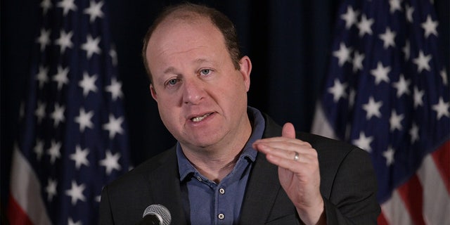 Jared Polis speaking with American flags in the background