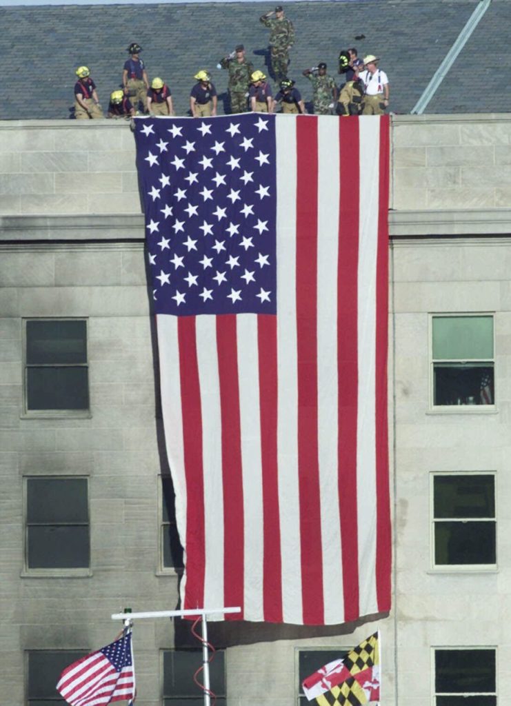 Firefighters unfurl an American flag from the roof of the Pentagon Wednesday, Sept. 12, 2001, as President Bush visits the area of the Pentagon where an airliner, hijacked by terrorists, crashed into the building on Tuesday. (AP Photo/Ron Edmonds)