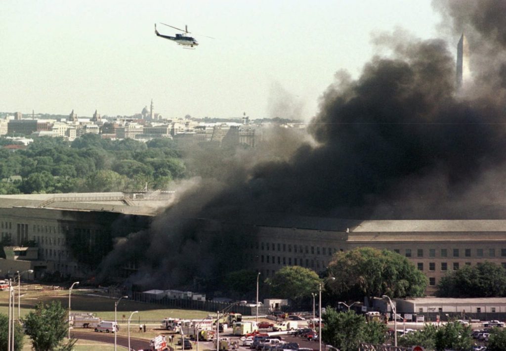 A helicopter flies over the burning Pentagon Tuesday, Sept. 11, 2001. The Washington Monument can be seen at right, through the smoke. The White House roof is visible in the trees of Washington at left. (AP Photo/Tom Horan)