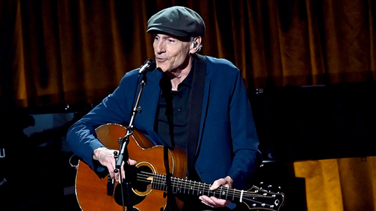 James Taylor wears a blue jacket, black shirt and pants and plays the guitar wearing a cap at the Gershwin Prize for Joni Mitchell