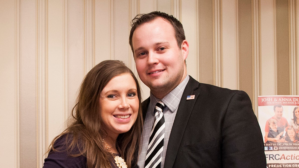 Anna Duggar wearing a dark purple dress standing next to her husband Josh Duggar in a dark suit and striped tie