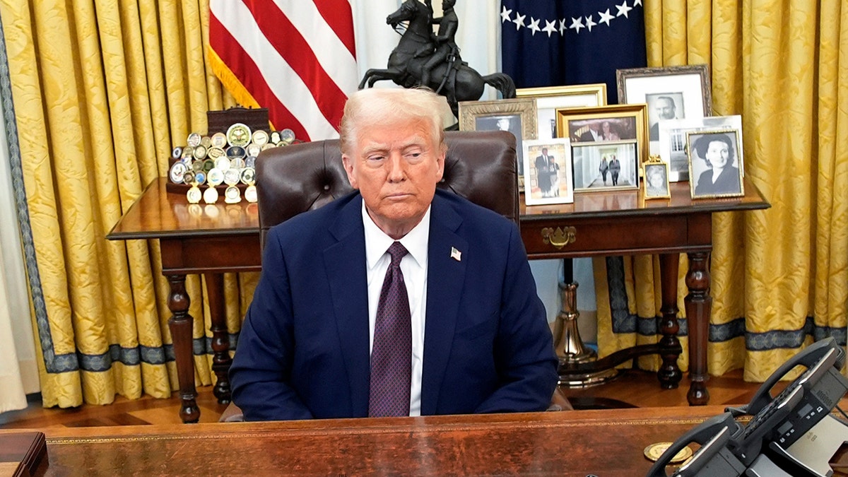 President Trump sits at the Resolute Desk with family photos behind him.