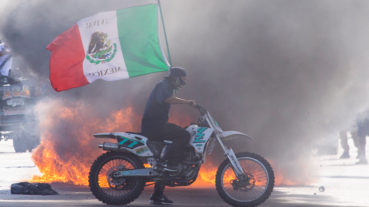 Rioter in LA on dirt bike holding Mexican flag, fire behind him