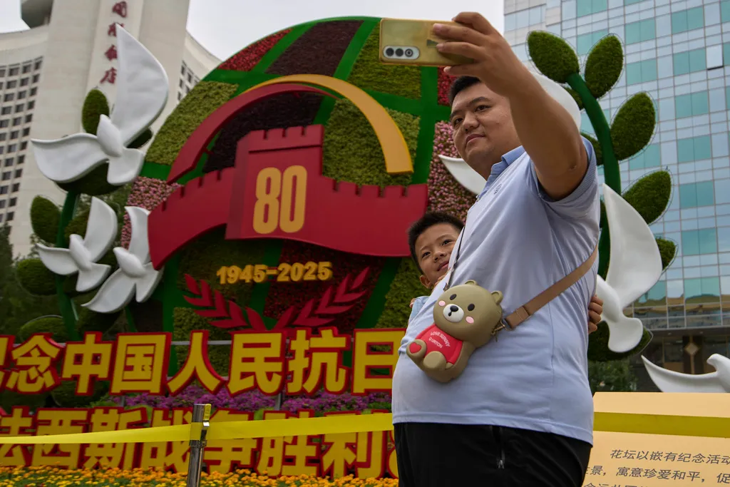 A man takes a selfie with a child at a floral decoration displaying a symbol of the 80th anniversary of the end of World War II.