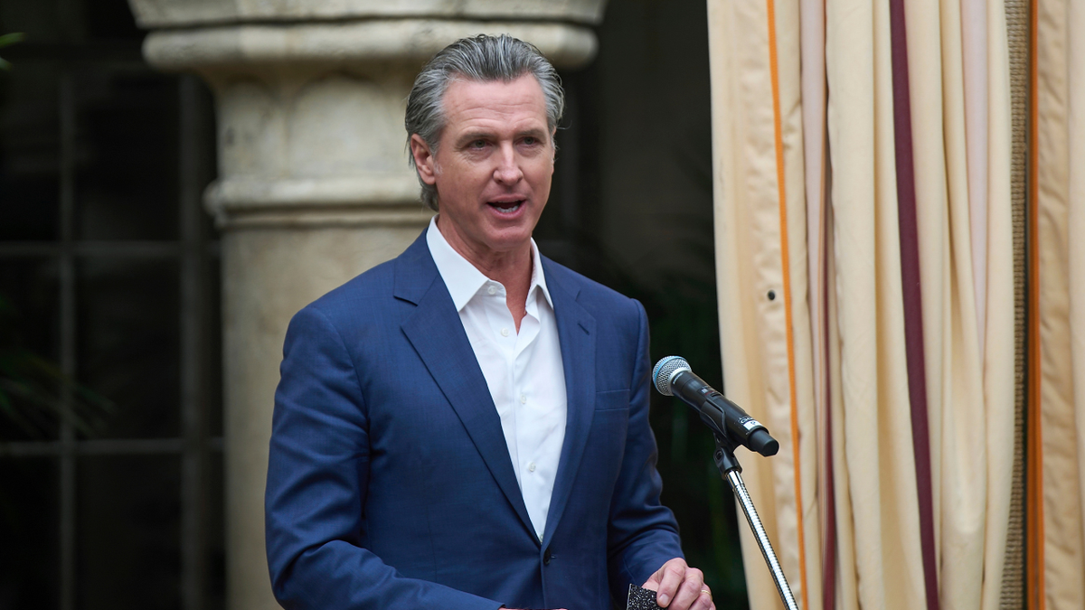 Gavin Newsom stands behind a podium wearing a navy suit and white button up shirt.