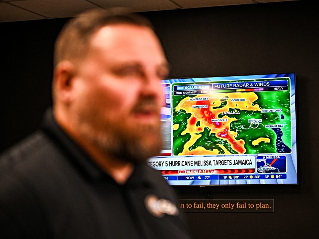 A weather news channel displays Hurricane Melissa updates as Billy Richardson, Director of US warehousing of Global Empowerment Mission, speaks during an interview with AFP at Global Empowerment Mission Headquarters in Miami, Florida, on October 27, 2024. Hurricane Melissa threatened Jamaica with potentially deadly rains after rapidly intensifying into a top-level Category 5 storm, as residents scrambled for shelter from what could be the island's most violent weather on record. Melissa has already been blamed for at least four deaths in Haiti and the Dominican Republic, and was set to unleash torrential rains on parts of Jamaica in a direct hit on the Caribbean island. (Photo by CHANDAN KHANNA / AFP)