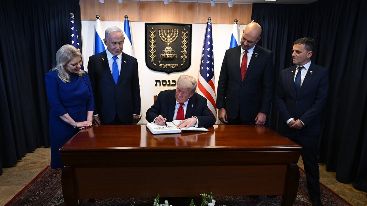 Donald Trump sitting at desk at the Knesset as he signs a guestbook with Israeli Prime Minister Benjamin Netanyahu and others watching.