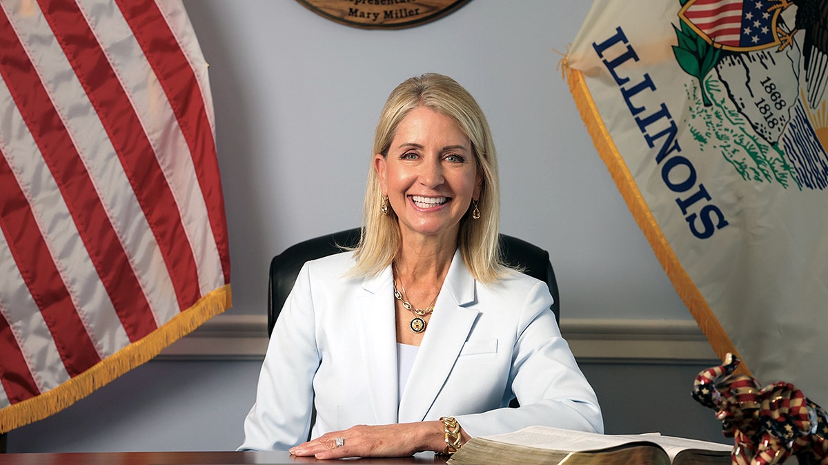 Rep. Mary Miller sits smiling at her desk with U.S. and Illinois flags behind her.