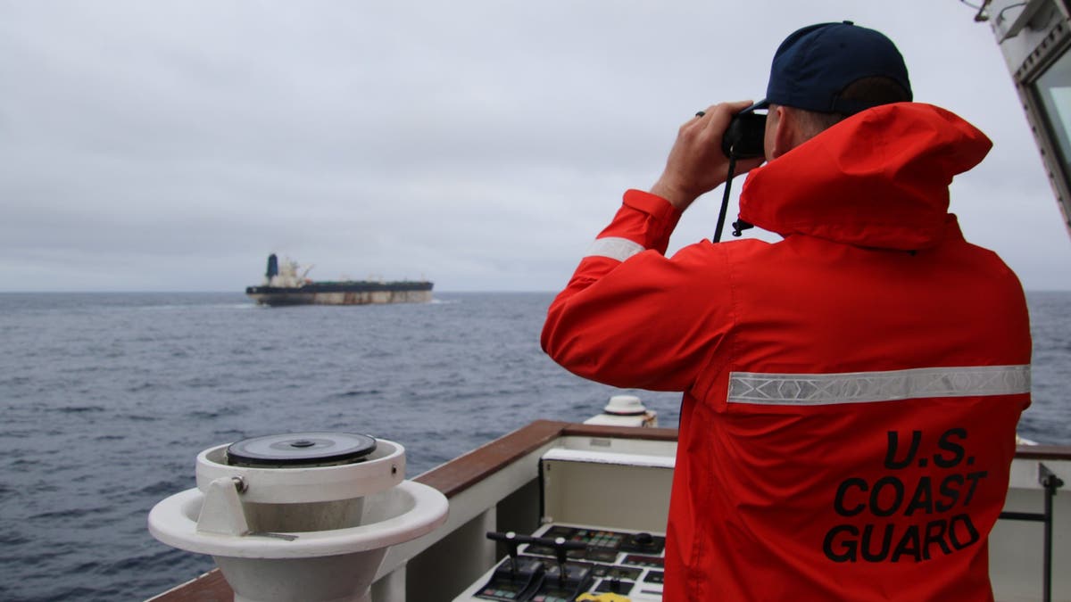 A member of the U.S. Coast Guard looking through binoculars at an oil tanker