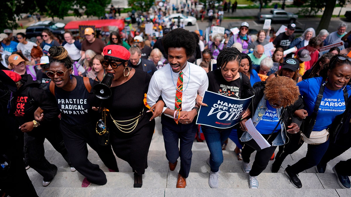Justin J. Pearson marches arm-in-arm with a crowd of protesters up stone steps while holding a megaphone.