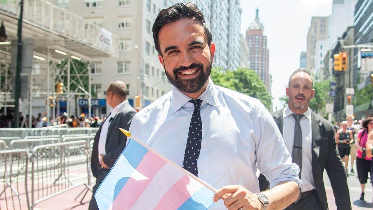 Zohran Mamdani smiles while holding a transgender pride flag in 2025