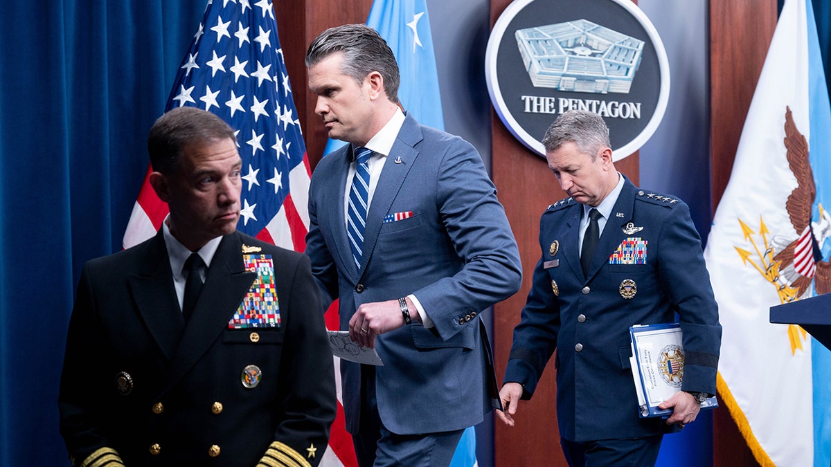Pete Hegseth, Gen. Dan Caine, and Adm. Brad Cooper walking away from a podium in a briefing room.