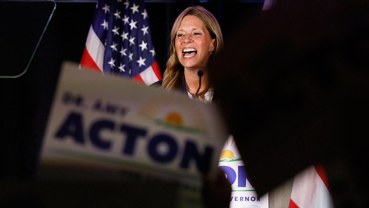 Dr. Amy Acton speaks at a podium with a wide smile in front of an American flag.